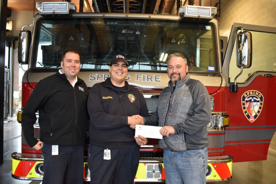 Three men stand in front of a fire truck, with two of them shaking hands while holding a document or check.