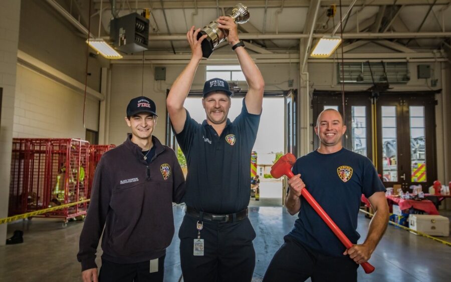 Three firefighters stand together in a fire station; the center firefighter raises a trophy, while the firefighter on the right holds a large red axe.