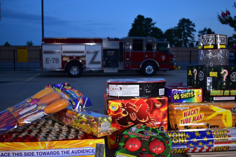 A variety of fireworks are displayed on a table in the foreground, with a red fire truck parked in the background.