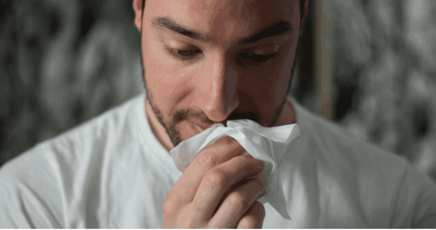 A man in a white shirt holds a tissue to his nose, appearing to sneeze or wipe his nose.