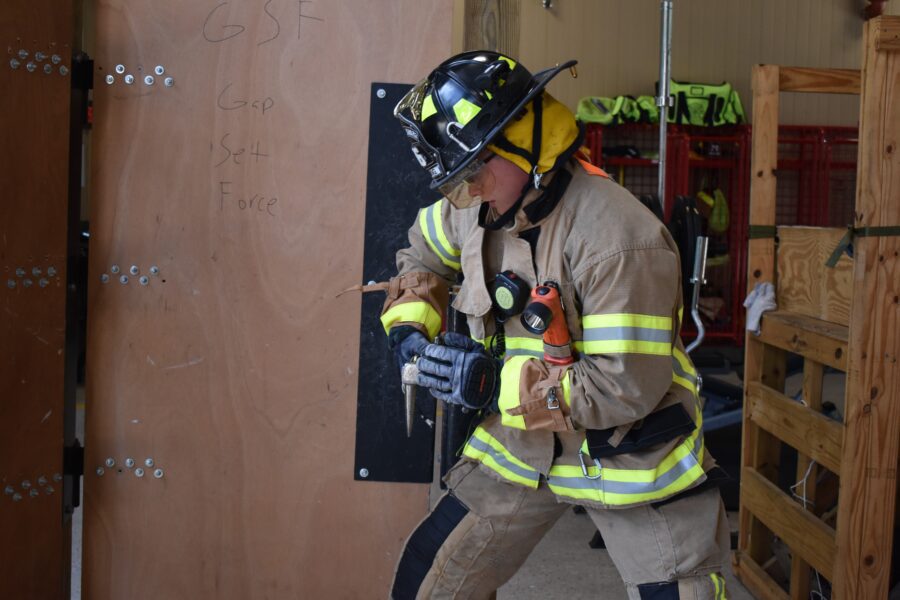 A firefighter in full gear uses a tool to force open a wooden door during a training exercise indoors.