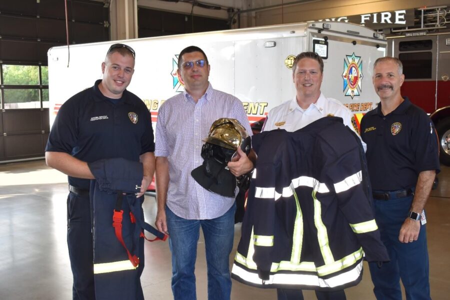 Four men stand inside a fire station; two are in dark uniforms, one in a white uniform holds fire gear, and one in casual clothes, with a fire truck in the background.