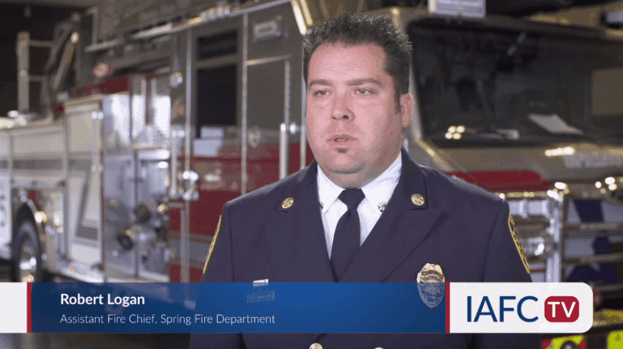 A man in a fire department uniform stands in front of fire trucks. Text overlay identifies him as Robert Logan, Assistant Fire Chief, Spring Fire Department. IAFC TV logo is visible.