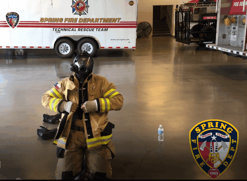 A firefighter in full gear kneels indoors by a water bottle, with a Spring Fire Department rescue vehicle visible in the background.