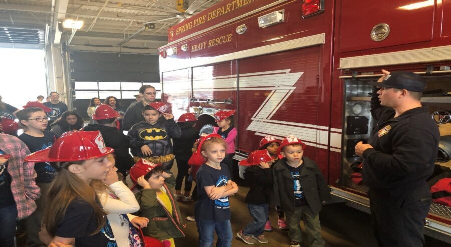 A firefighter speaks to a group of children and adults wearing red toy helmets, standing next to a fire truck inside a fire station.