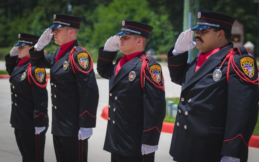 Four uniformed firefighters stand in a row outdoors, saluting with their right hands, wearing black dress uniforms and hats with department patches visible on their sleeves.