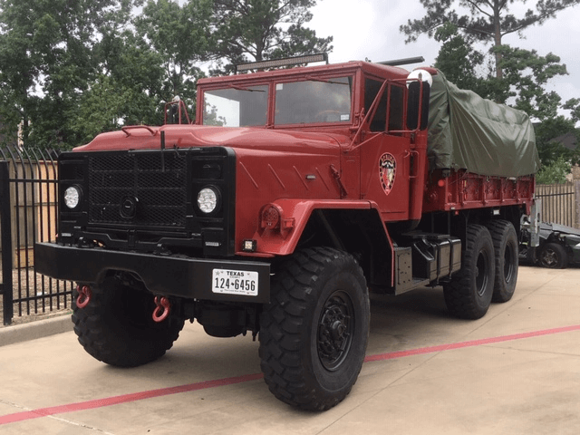 A large red military-style truck with six wheels and a covered cargo area is parked on a concrete surface near a black metal fence.