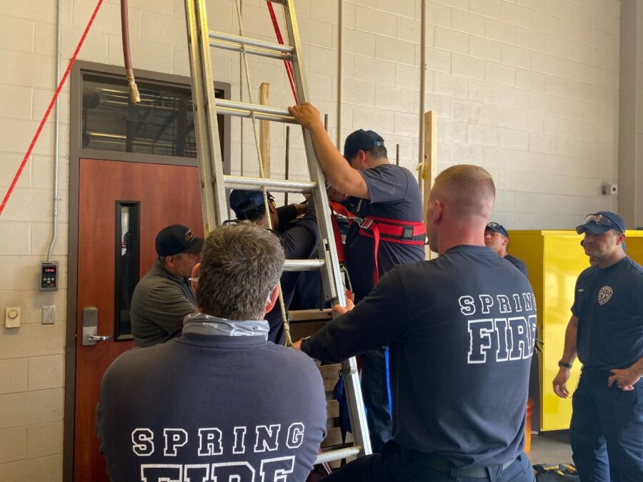Several firefighters, wearing "Spring Fire" uniforms, practice a rescue drill indoors using a ladder and safety harness near a door and yellow cabinet.