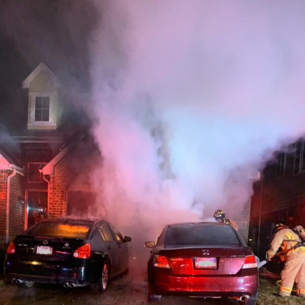Two firefighters respond to heavy smoke rising from two parked cars in front of a brick house at night.