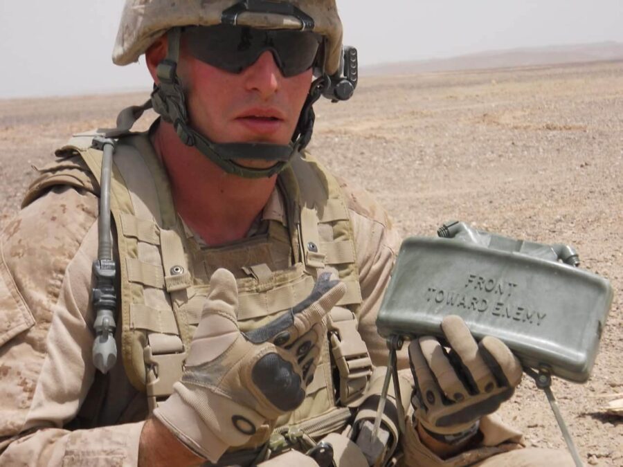 A soldier in desert camouflage gear holds a Claymore mine marked "Front Toward Enemy" while sitting on sandy, rocky terrain.