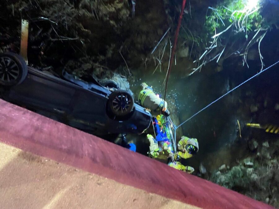 Firefighters rescue a person from a car overturned in a creek at night, with emergency ropes and equipment visible.