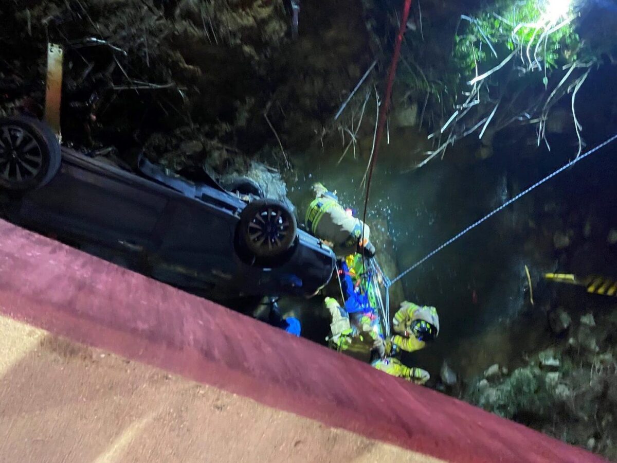 Firefighters rescue a person from a car overturned in a creek at night, with emergency ropes and equipment visible.