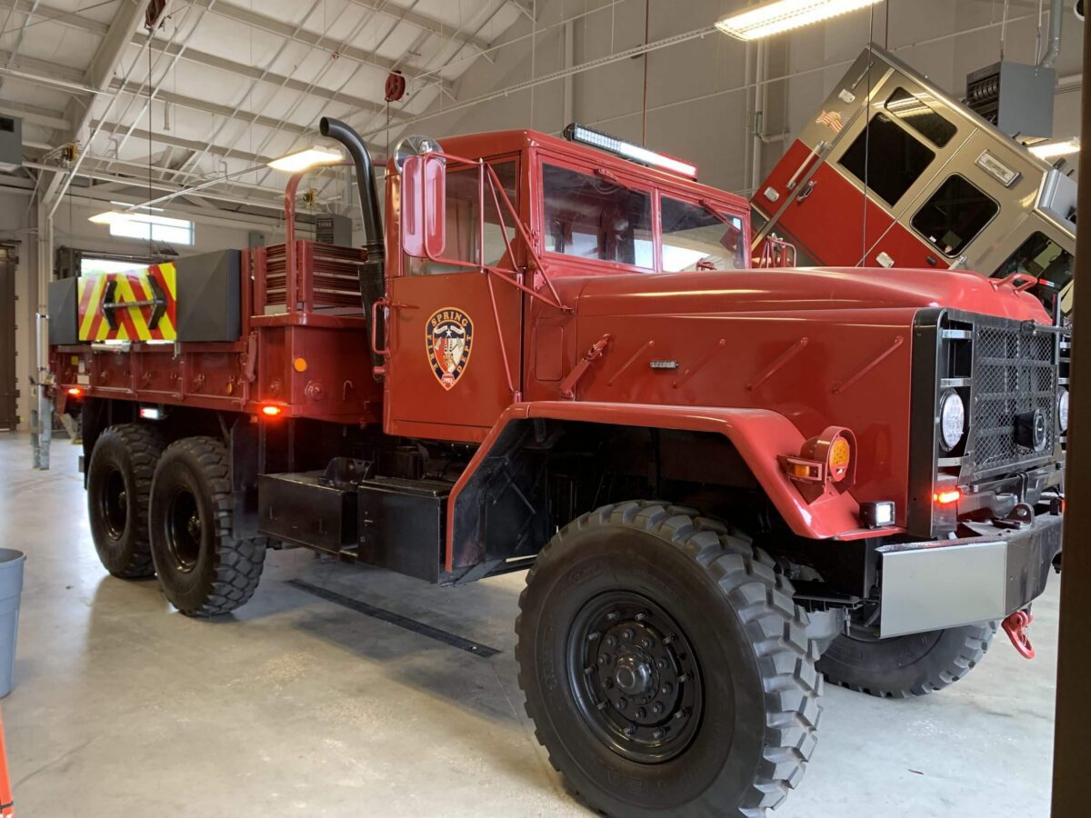 A large red fire department utility truck with six wheels is parked inside a fire station garage; other emergency vehicles are visible in the background.