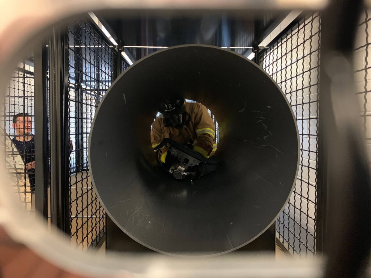 A firefighter crawls through a large metal pipe during a training exercise as two people observe from outside.