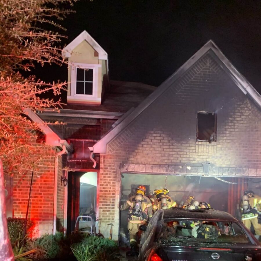 Firefighters respond to a house fire at night; a burnt car is parked in the driveway, and smoke damage is visible on the garage and upper windows.