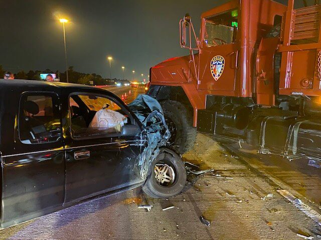 A black car with a deployed airbag has crashed head-on into a large red emergency vehicle at night on a highway, with visible front-end damage to the car.