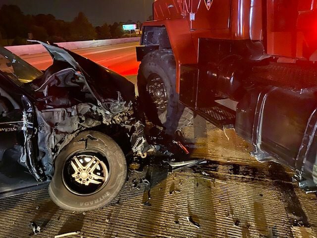 A black car with severe front-end damage is crashed into the side of a large red truck on a highway at night.