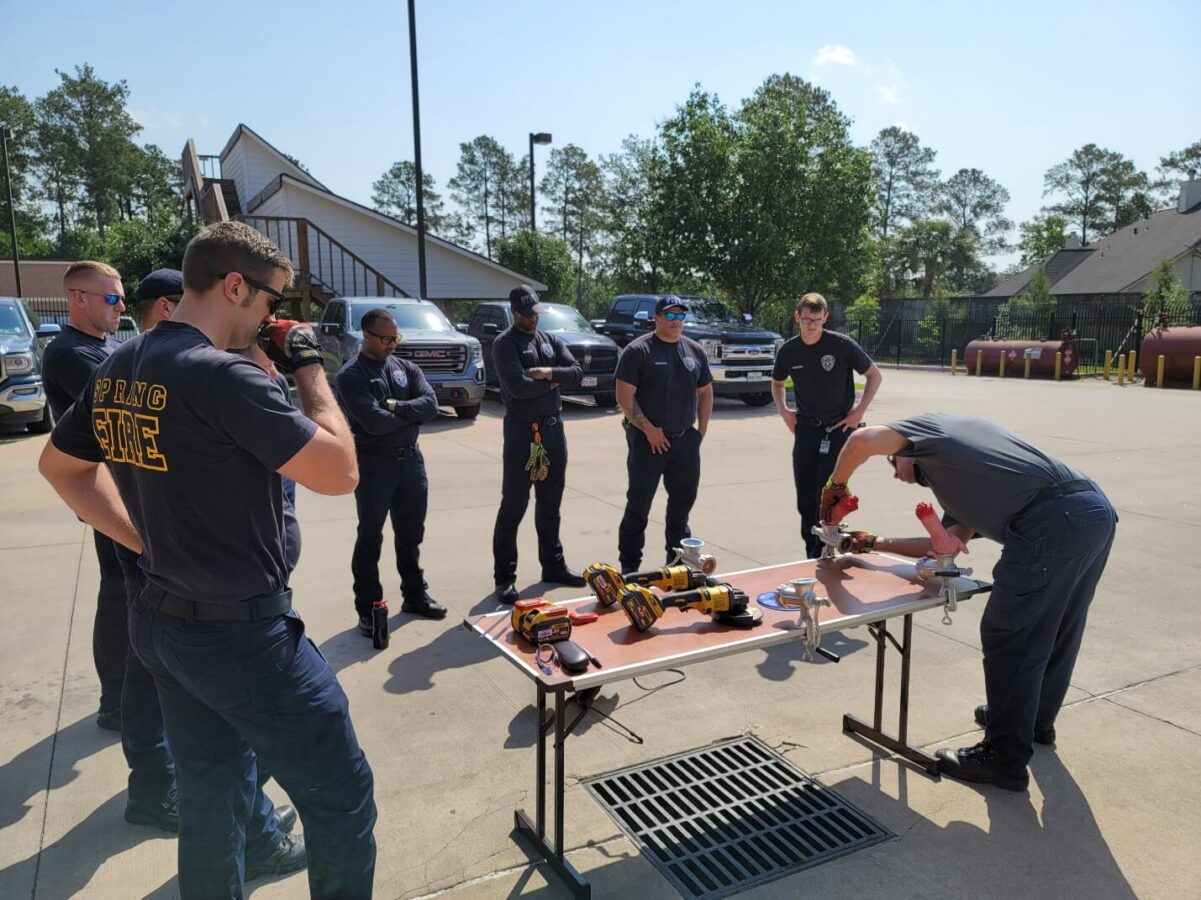 Several firefighters stand around two tables outdoors, observing a demonstration of rescue tools and equipment by a person in uniform.