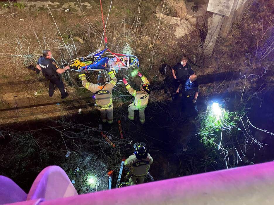 Emergency responders use a stretcher to rescue a person from a wooded, rocky area at night, illuminated by flashlights and vehicle lights.