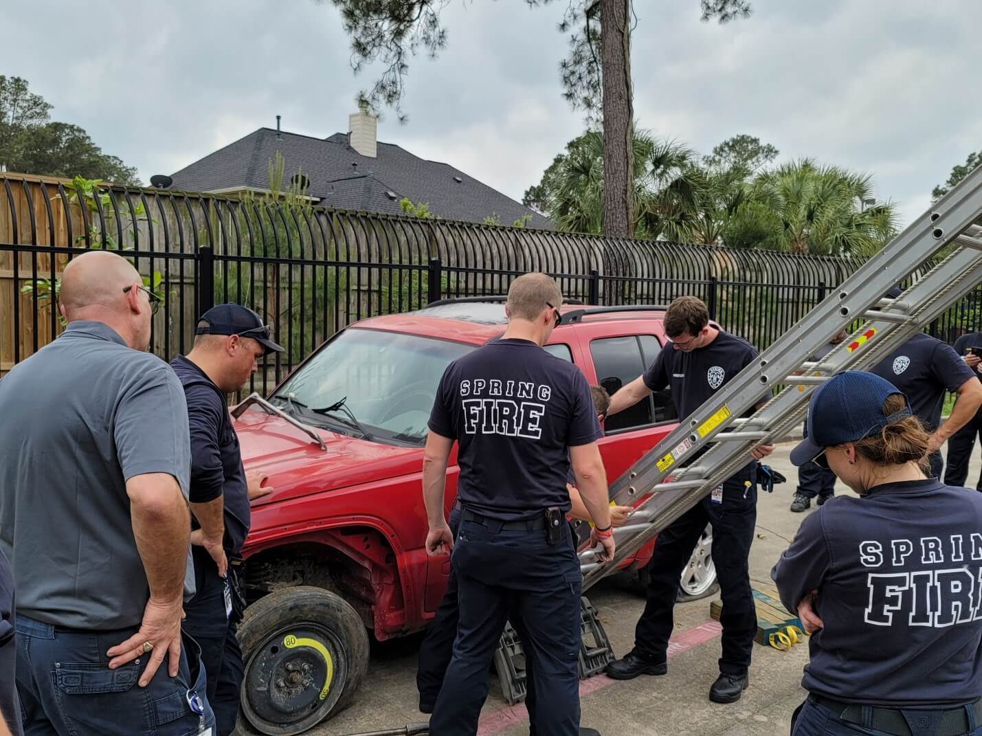 Firefighters in uniform gather around a red SUV with a missing front wheel, using tools and a ladder in a residential area.