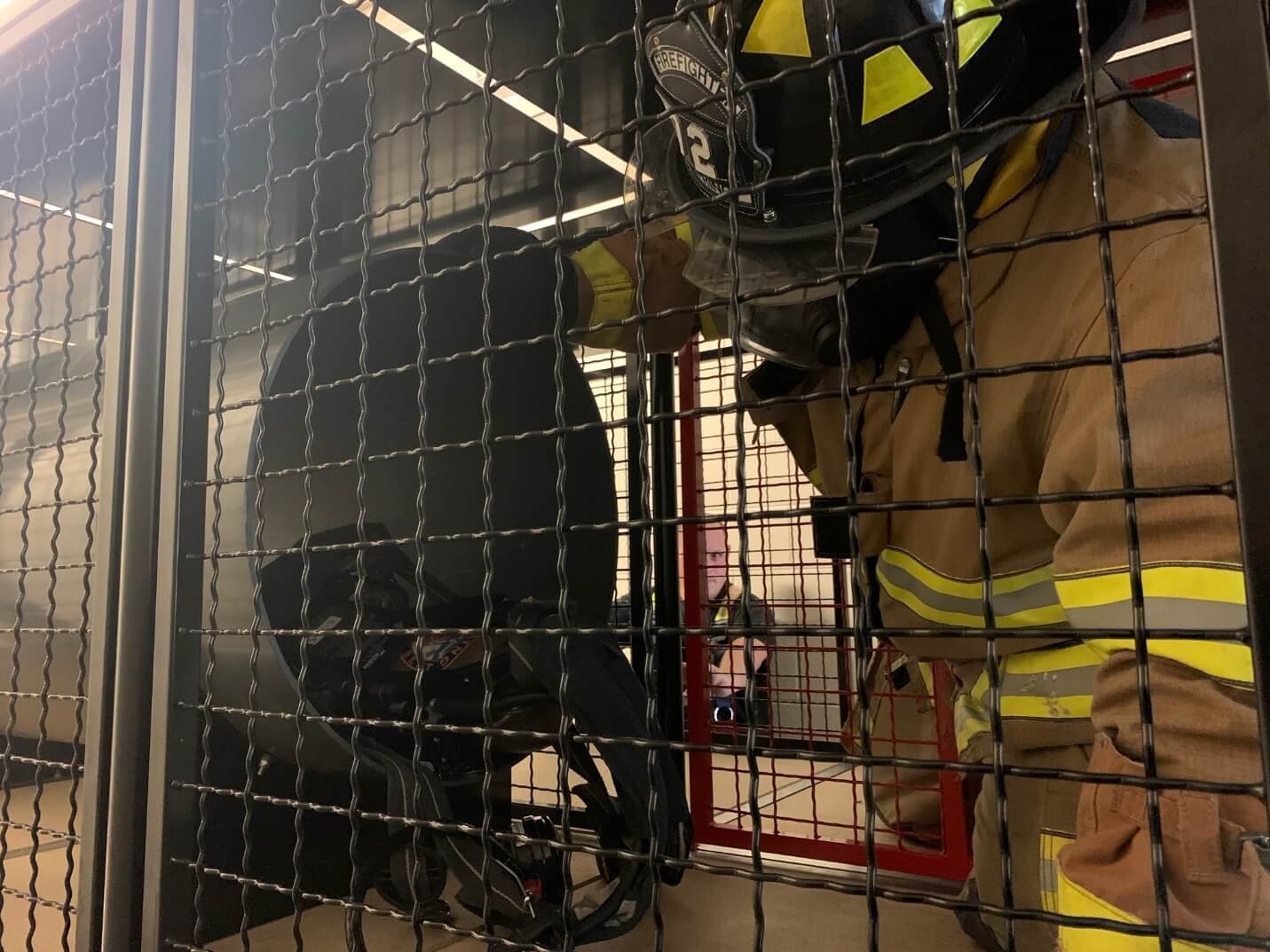 A firefighter in full gear stands next to a black helmet stored inside a metal mesh locker.