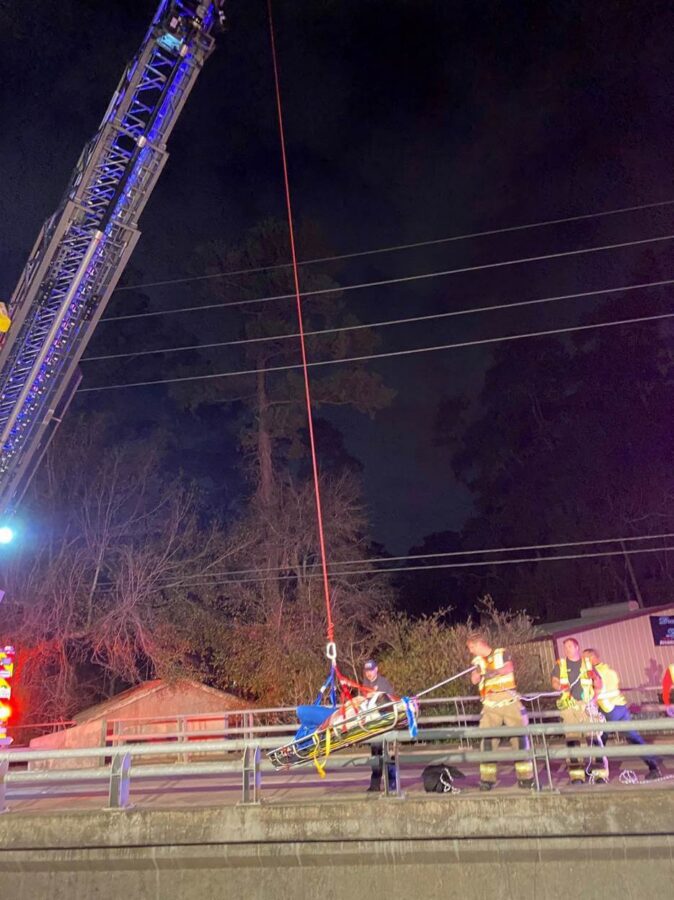 Firefighters use a crane and stretcher to perform a nighttime rescue on a bridge, with power lines and trees visible in the background.