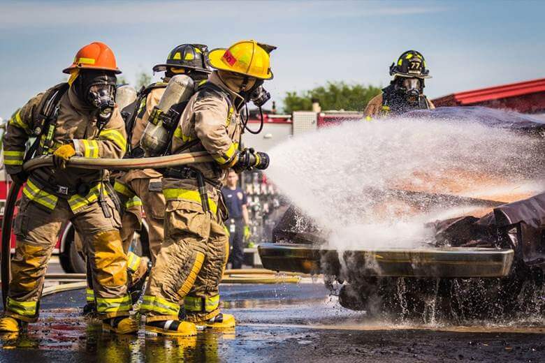 Firefighters in full gear spray water onto a burning vehicle during a daytime emergency response.