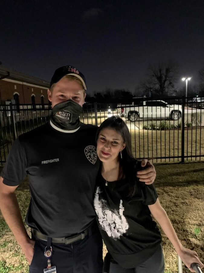 A firefighter in uniform and mask stands next to a woman dressed in casual clothes; they are outside by a fence at night.