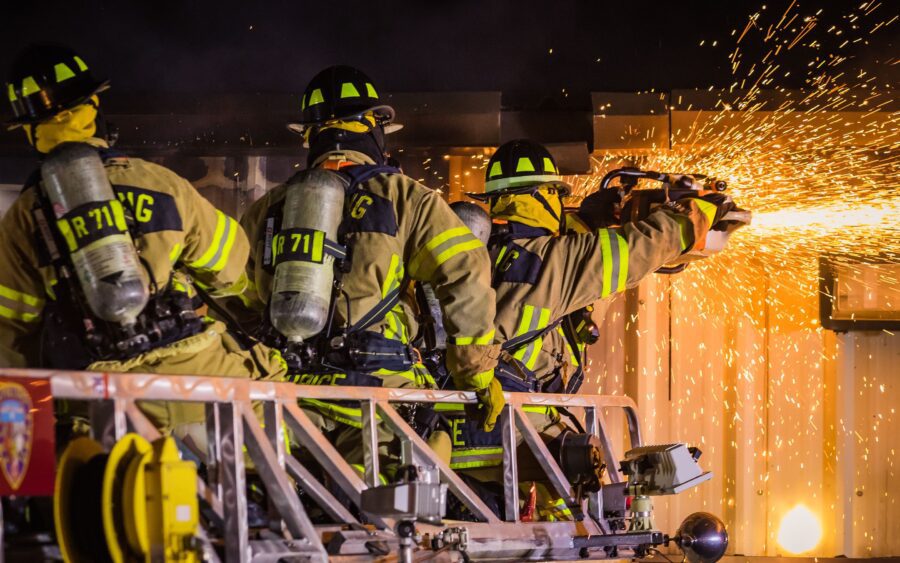 Three firefighters in full gear cut through a metal structure with a power saw, producing a shower of sparks during a nighttime operation.