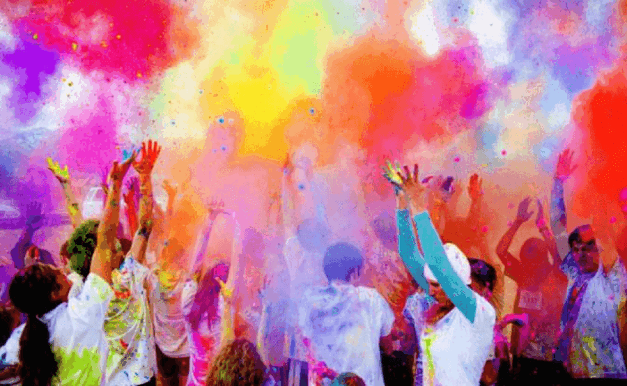 A crowd of people wearing white clothes throw colorful powder into the air during a festival or celebration.