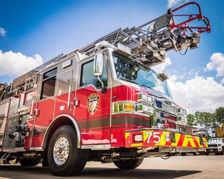 A red fire truck with a ladder on top is parked outdoors under a blue sky with some clouds. The vehicle is labeled "Spring Fire" and numbered 75.