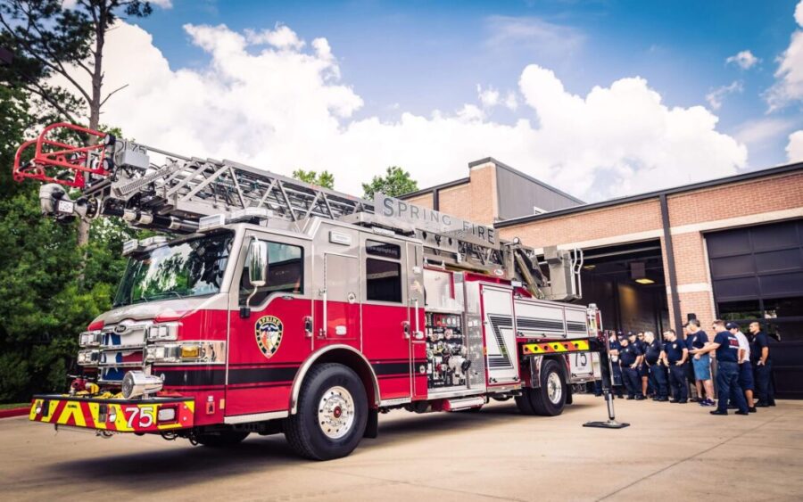 A red fire truck with a ladder is parked outside a fire station, while a group of firefighters stands together near the building.