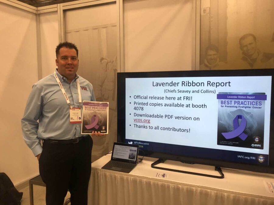 A man stands next to a display featuring the Lavender Ribbon Report on best practices for preventing firefighter cancer, holding a copy of the report beside a monitor with report details.