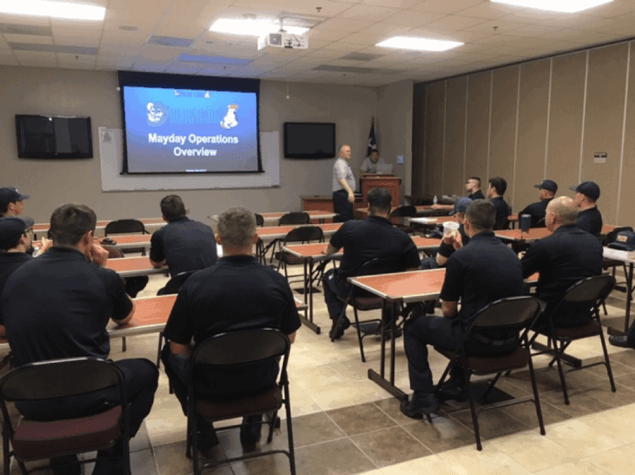 A group of uniformed individuals attends a classroom presentation titled "Mayday Operations Overview," with a speaker standing at the front near a screen.