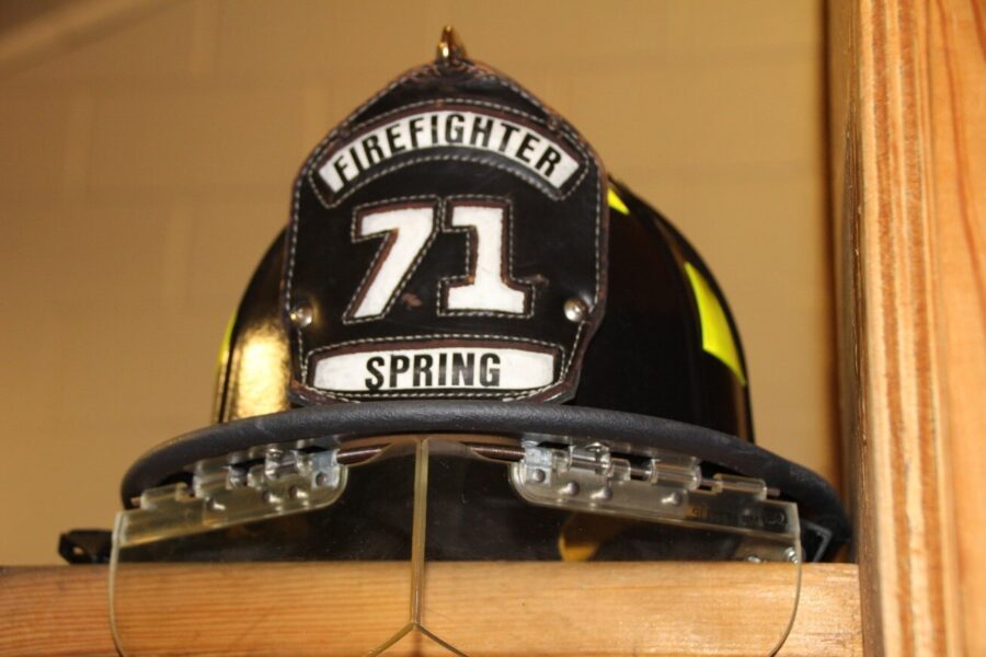 A black firefighter helmet labeled "Firefighter 71 Spring" rests on a wooden shelf with protective goggles attached.