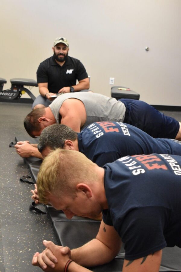 Four men hold a plank position on mats in a gym while another man, wearing a cap and holding a clipboard, observes from behind.