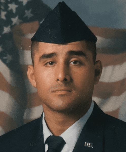 A man wearing a U.S. Air Force dress uniform and cap poses for a formal portrait in front of an American flag backdrop.