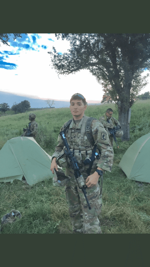 A soldier in camouflage uniform stands in front of green tents with two other soldiers in the background on a grassy field near a tree.