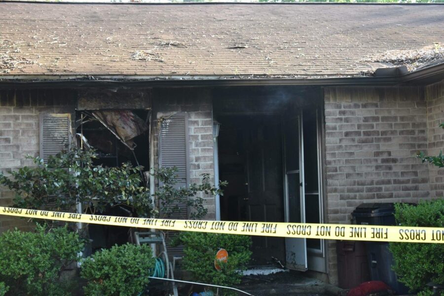 A brick house with a fire-damaged window and door, surrounded by "fire line do not cross" tape, showing burn marks and debris.