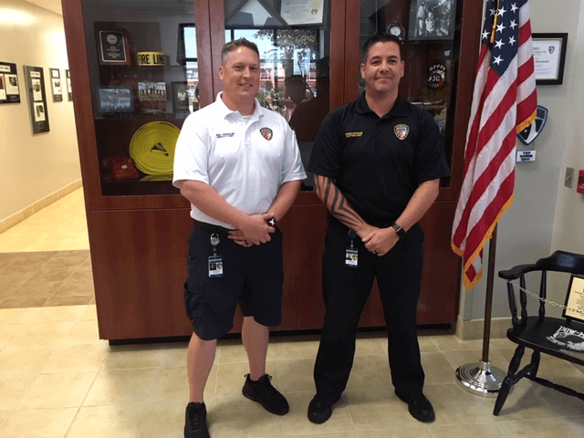 Two uniformed men stand side by side indoors, one in a white shirt and shorts, the other in a black shirt and pants, with a US flag and display case in the background.