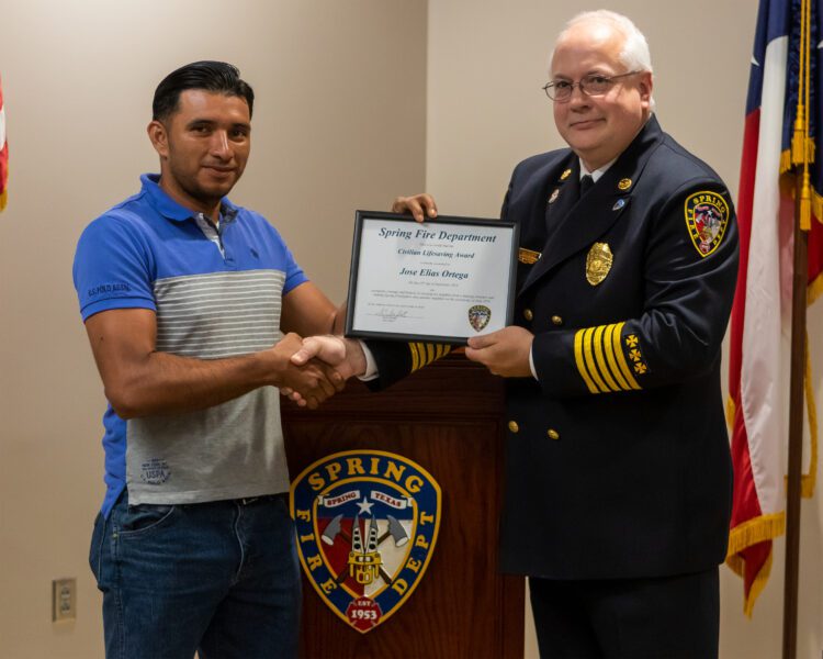 A man in a blue shirt receives a certificate from a uniformed fire chief while shaking hands in front of a Spring Fire Department sign.