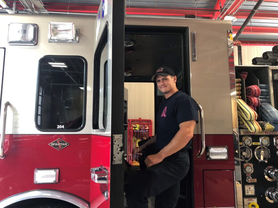 A firefighter in uniform stands on the step of a fire truck, holding equipment, with hoses and gauges visible on the side.