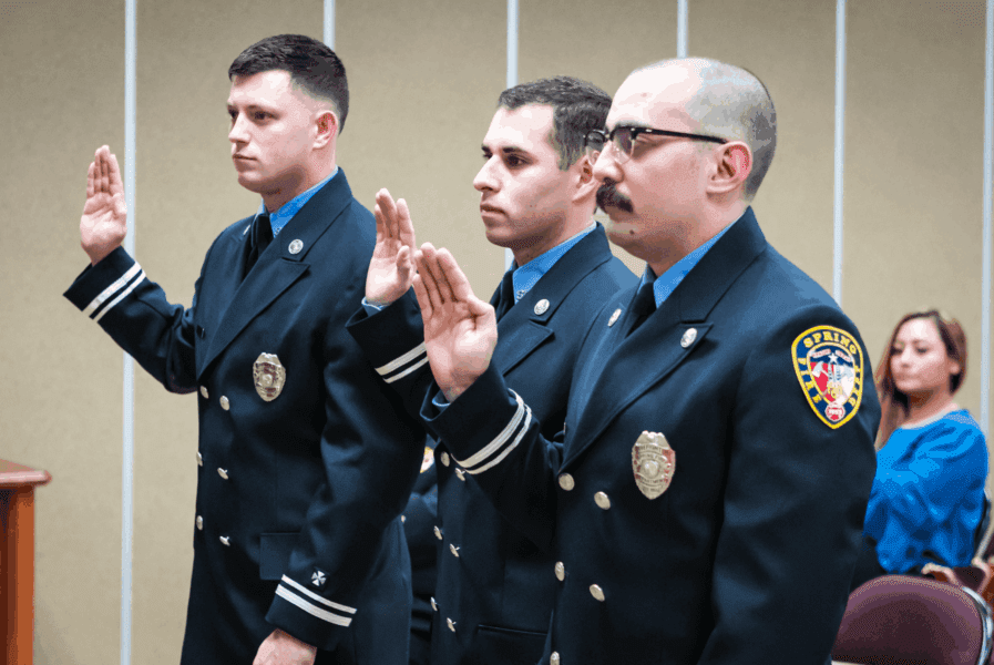 Three firefighters in uniform stand with their right hands raised, taking an oath in a formal indoor setting, with a seated woman in the background.