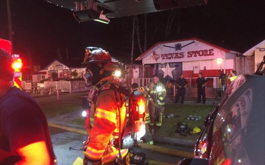 Firefighters in full gear respond at night outside a building labeled "Texas Store," with emergency vehicles and other personnel present.