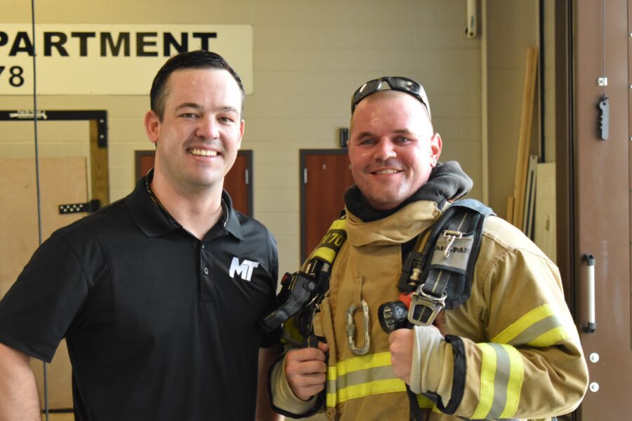 Two men pose indoors; one wears a black polo shirt with an "MT" logo, the other is dressed in firefighter gear, standing beside each other and smiling at the camera.