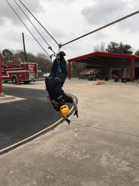 A person in safety gear hangs upside down on a zip line near a fire station, with a fire truck and people visible in the background.