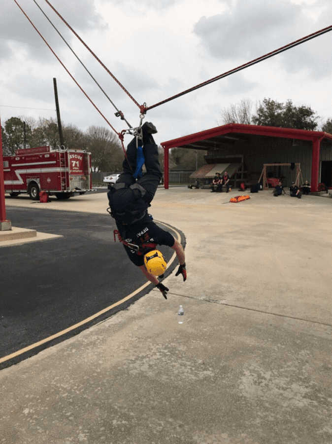 A person in safety gear hangs upside down on a zip line near a fire station, with a fire truck and people visible in the background.