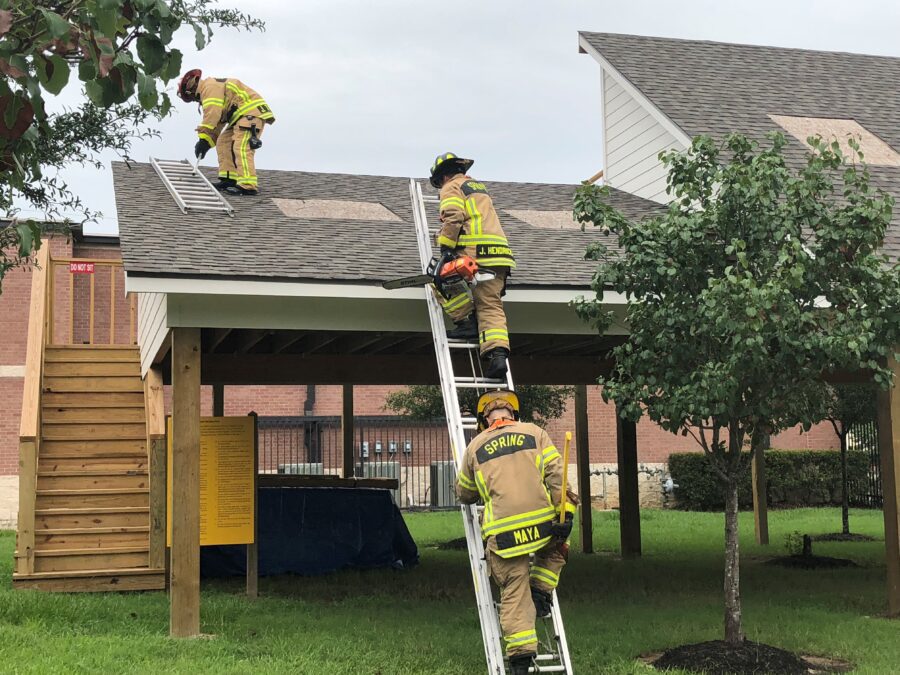 Three firefighters in gear practice roof ventilation techniques on a training structure, using ladders and tools, with trees and grass in the background.