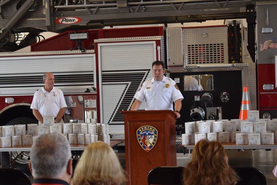 A fire chief speaks at a podium in front of a fire truck, with another firefighter and tables stacked with boxes nearby, while an audience listens.