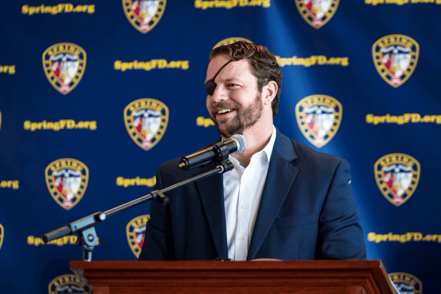 A man in a suit with an eye patch speaks at a podium, with a microphone and a backdrop displaying SpringPD.org and police badges.